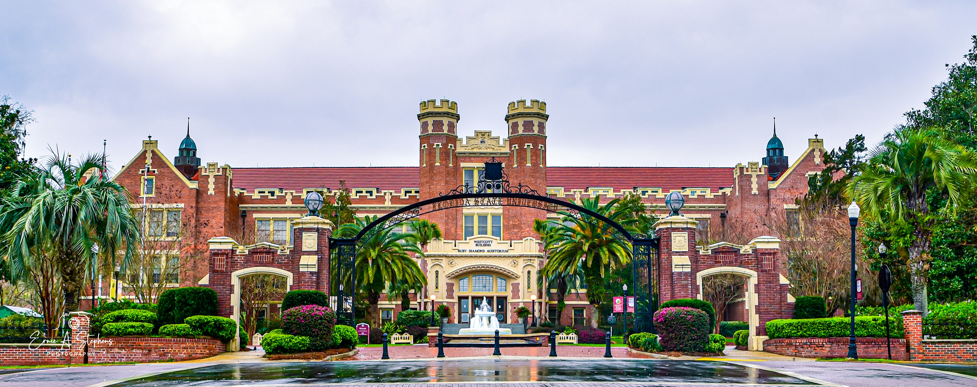 An image of a long, two-story red brick building with two small towers in the middle. The building has palm trees at both ends, as well as to either side of the arched front door. In front of the building is an arched iron gate with &quot;Florida State University&quot; spelled out on it.