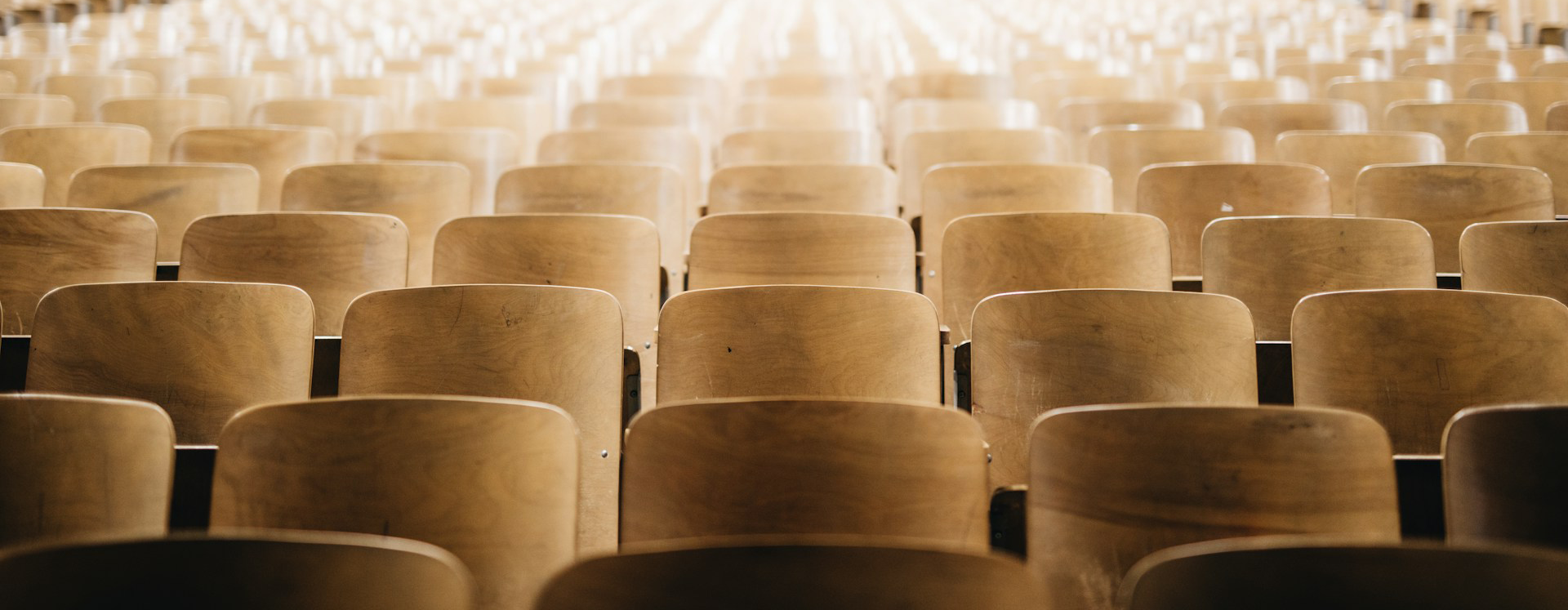 A photo of empty chairs in an auditorium. The chairs have simple, pale wooden backs. Photo is by Nathan Dumlao, from Unsplash.