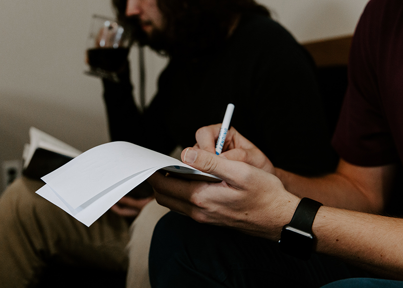 A man takes notes on paper next to another man with long hair who is drinking coffee.