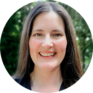 A woman with long brown hair, parted in the middle, smiles at the camera. She's standing in front of a hedge or tree with green leaves.