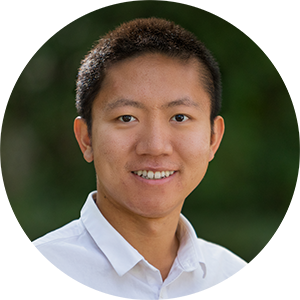 A young man smiles at the camera. He has very short black hair and wears a white collared shirt with the top button open.