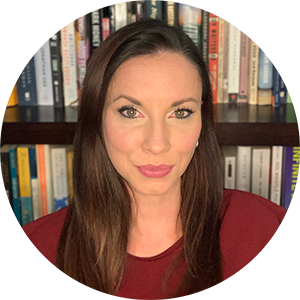 A woman with long, straight brown hair looks at the camera. She is wearing a dark red shirt, and is in front of a bookshelf full of books.