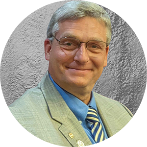 An older man wearing glasses smiles at the camera. He is wearing a light green suit jacket, a blue shirt, and a yellow and blue striped tie.