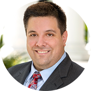 A man smiles widely at the camera. He has short brown hair and wears a charcoal suit, a light blue shirt, and a colorful tie.