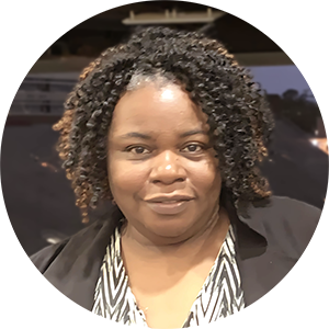 A Black woman with shoulder-length hair looks at the camera. She is wearing a black jacket with lapels and a black and white striped shirt.