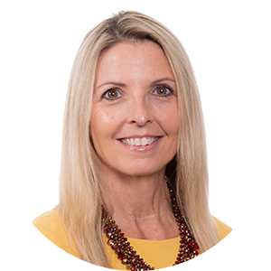 A white woman smiles at the camera. She has straight blonde hair that reaches to her shoulders; she wears a yellow collarless blouse and a multi-string necklace.