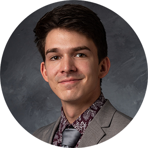 A man with short dark hair looks at the camera. He is wearing a grey suit and tie, and a maroon collared shirt with white floral outlines on it.