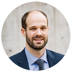 ​A white man with a short beard smiles at the camera. He is wearing a navy suit and tie with a blue shirt.