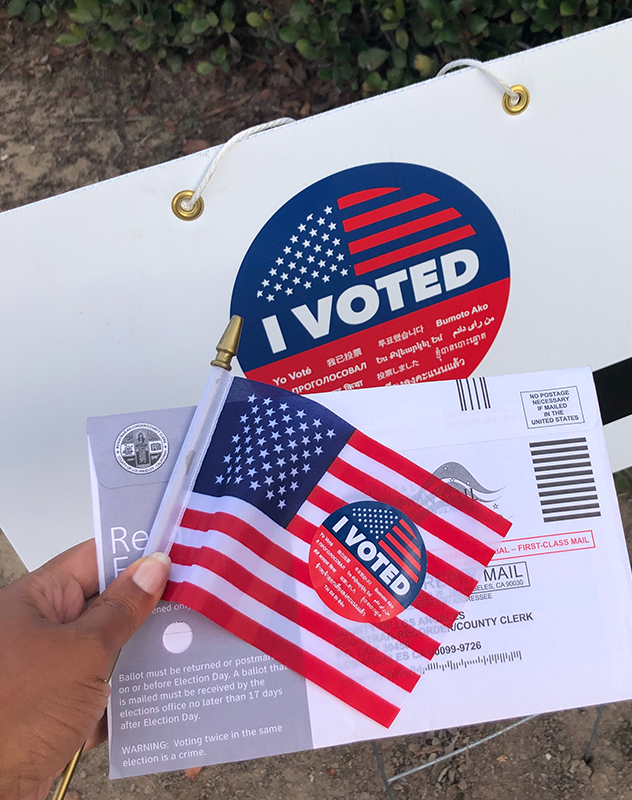 A woman with brown skin and long nails holds a miniature American flag along with an absentee ballot and an "I Voted" sticker.