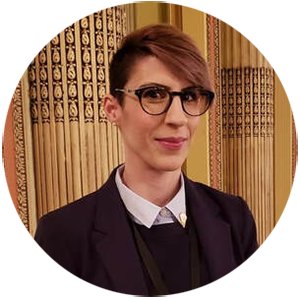 A woman with short brown hair and glasses smiles at the camera. She is wearing a suit jacket and dark vest over a button-up shirt.