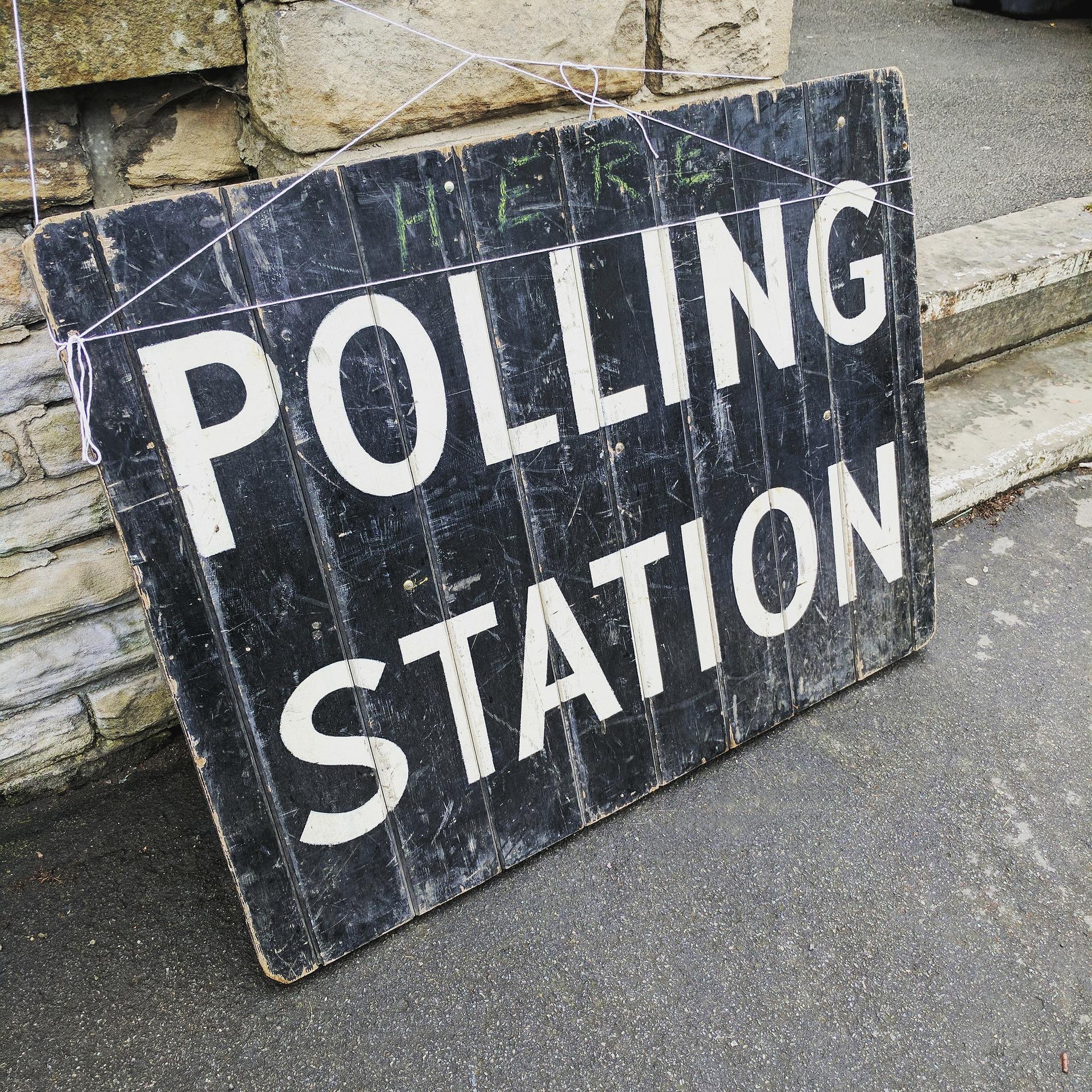 A black sign leans against a brown stone wall. The sign reads "Polling Station" in white all caps lettering.