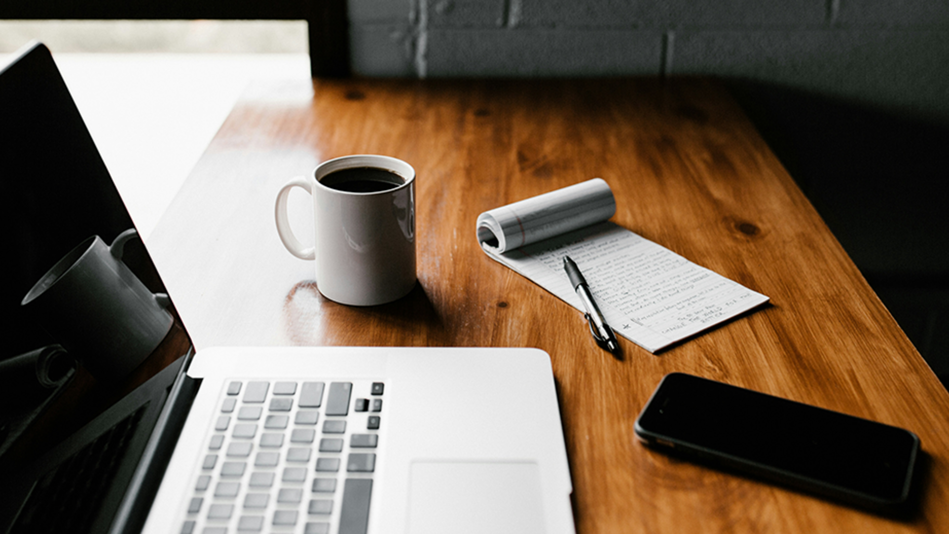 A photo of a laptop on a wooden table, in front of a window. There is a phone in the foreground, and a full mug of coffee in the background.