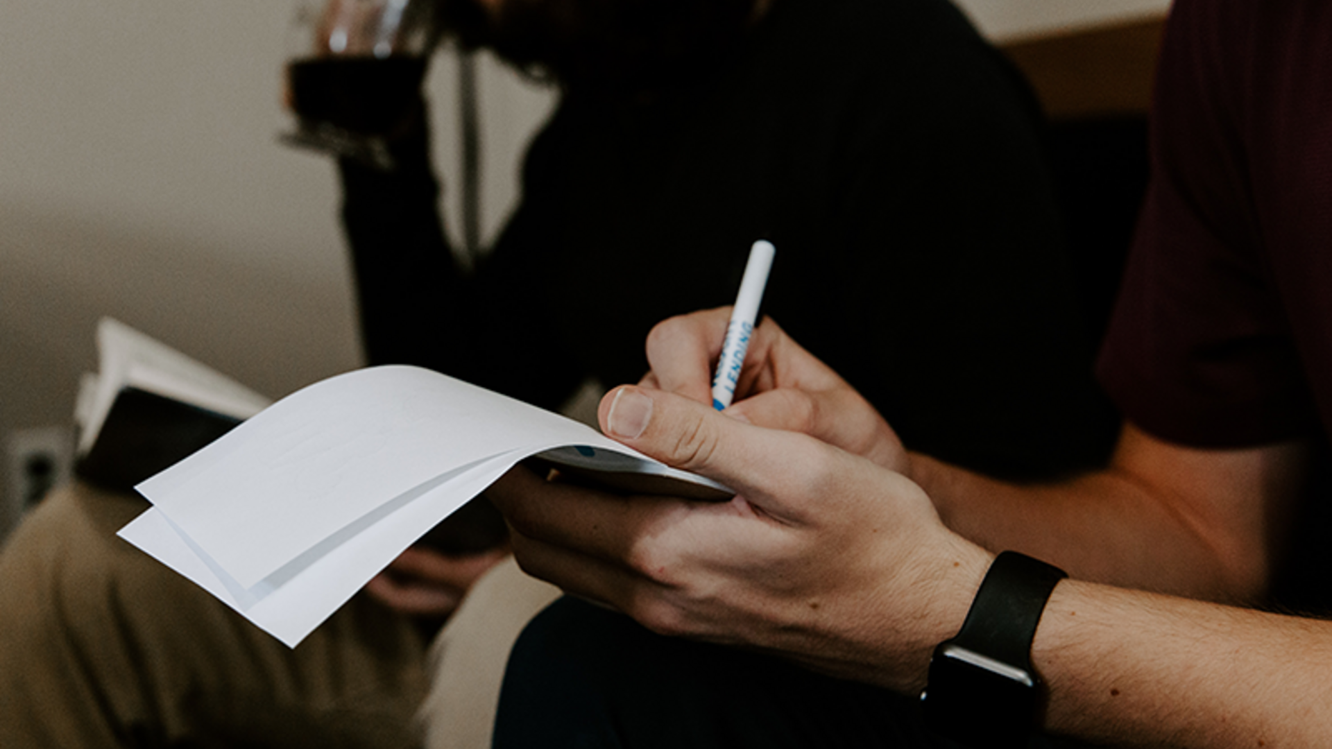 A man takes notes on paper next to another man with long hair who is drinking coffee.