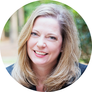 A woman with shoulder-length blonde hair smiles at the camera. She is wearing a dark suit jacket, and we can see green trees in the background behind her.