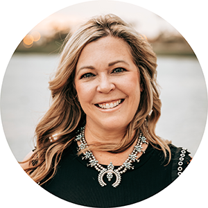 A woman with wavy blonde hair smiles; she is wearing an intricate necklace and a black sleeveless top with white decorative eyelets along the edges.