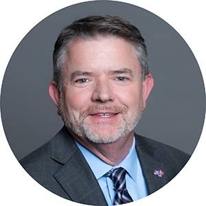 A white man with salt and pepper hair and a short beard smiles at the camera. He is wearing a grey suit with a lapel pin, a light blue collared shirt, and a blue diagonally striped tie.