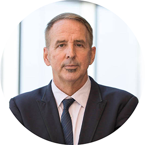 A man with short hair looks at the camera. He is wearing a dark suit coat and a faintly striped collared shirt with a dark tie.